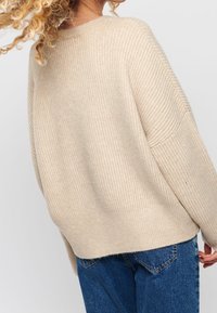 Beige žebrovaný svetr s uvolněným střihem a spadlými rameny. Vyrobený z měkkého materiálu, s kulatým výstřihem a mírně zakřiveným lemem.