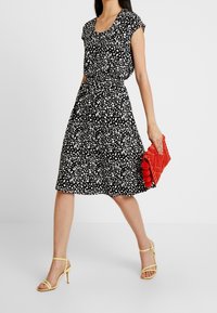 Robe noire et blanche à pois avec des manches courtes, taille froncée, jupe fluide et une pochette rouge texturée ; associée à des sandales jaunes à brides.