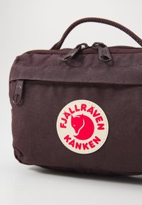 Pruun Fjällräven Kånken kott, millel on ümmargune logo, varustatud lukuga ja tekstuurse kangaga, kompaktne disain koos ülemise käepidemega mugavaks kandmiseks.