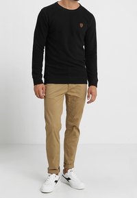 Mężczyzna ubrany w czarną koszulę z długim rękawem, przewinięte khaki spodnie i białe trampki stoi na tle jednolitego białego tła.