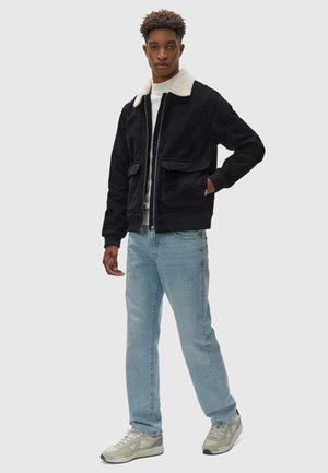 Black corduroy jacket with a cream shearling collar, front zip, and two large chest pockets, paired with light blue jeans and gray sneakers.