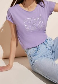 Nainen istuu laventelinvärisessä t-paidassa, jossa on hopeateksti "luonnolliset VÄRIMALLIT", ja vaaleansinisten korkeavyötäröisten farkkujen kanssa, joissa on pieniä kulumia.