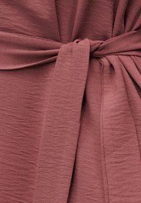 Κοντινό πλάνο υφάσματος σε αποχρώσεις σκόνης τριαντάφυλλου δεμένου με έναν απλό κόμπο, που αναδεικνύει την υφή του υλικού και τις απαλές πτυχές.