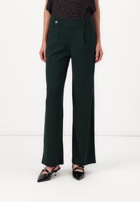 Groene wijde broek met twee frontplooien en decoratieve knopen op de tailleband, gecombineerd met zwarte pumps met spitse neuzen. Glad stoffen textuur.