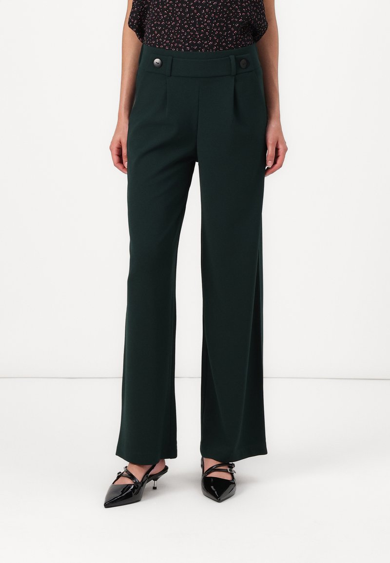 Groene wijde broek met twee frontplooien en decoratieve knopen op de tailleband, gecombineerd met zwarte pumps met spitse neuzen. Glad stoffen textuur.
