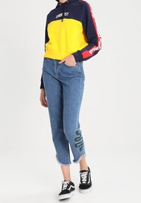 Felpa con cappuccio corta gialla e blu navy con coulisse, abbinata a jeans azzurri chiari con ricamo di un serpente verde. Scarpe da ginnastica nere e bianche.