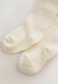 Calcetines de punto suaves de color crema con textura acanalada, acostados planos sobre una superficie blanca.