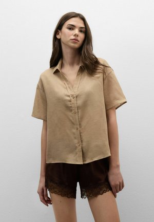 Junge Frau steht vor einer schlichten Wand, trägt ein beige kurzärmeliges Hemd mit Knöpfen und dunkelbraune Shorts mit Spitzenbesatz.