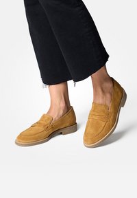 Sandfarbene Wildleder-Loren mit flacher Sohle, die ein gesticktes Design und zwei dekorative Bänder über der Vorderseite aufweisen. Getragen mit verkürzten schwarzen Hosen.