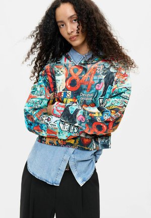 Ung kvinde med krøllet hår iført en farverig sweatshirt med katte, tal og tekst over en blå denims skjorte og sorte bukser.
