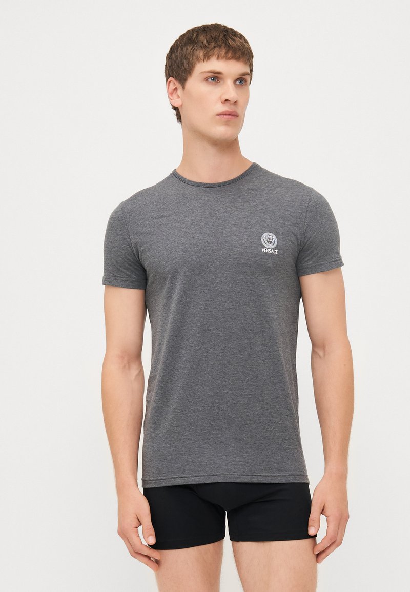 Graues T-Shirt mit kurzen Ärmeln und rundem Ausschnitt, das ein kleines Versace-Logo auf der Brust trägt. Das Material wirkt weich und leicht strukturiert.