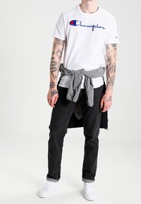 Camiseta blanca de manga corta con el logo "Champion" bordado en azul, combinada con jeans negros y un suéter gris anudado. Zapatillas blancas.