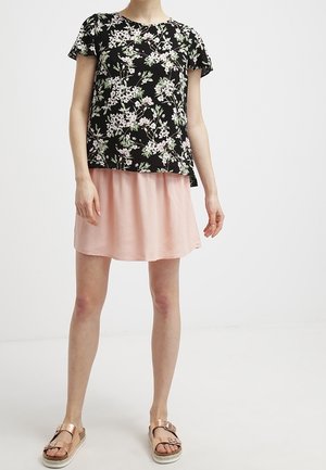 Femme portant un haut court à manches courtes noir à motifs floraux, une jupe rose clair et des sandales métalliques couleur or rose, debout sur un fond blanc.