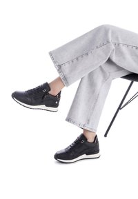 Zapatillas deportivas negras con cuero texturizado, diseño con cordones, suelas blancas y un sutil logo en la parte posterior. Combinadas con unos jeans de pierna ancha en gris claro.