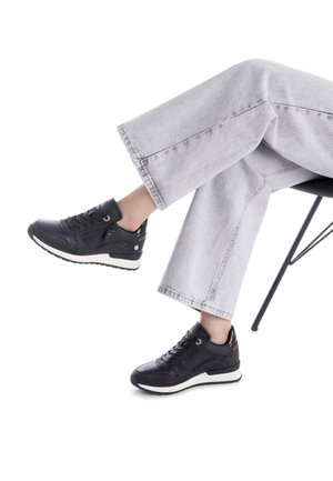 Zapatillas deportivas negras con cuero texturizado, diseño con cordones, suelas blancas y un sutil logo en la parte posterior. Combinadas con unos jeans de pierna ancha en gris claro.