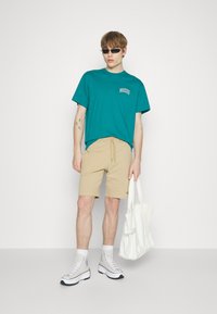 T-shirt di cotone teal con logo, pantaloni corti beige con cordoncino, borsa a tracolla bianca in tela e sneakers alte grigio chiaro con suole nere.