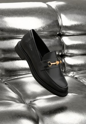 Mocassin en cuir noir à finition lisse, orné d'une ferrure dorée à l'avant, avec une semelle plate en caoutchouc, présenté sur un fond métallique.