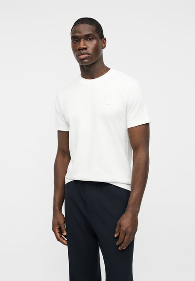 Άνδρας στέκεται φορώντας ένα απλό λευκό κοντομάνικο μπλουζάκι και σκούρο παντελόνι, μπροστά σε φωτεινό φόντο.