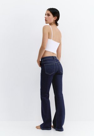 Frau mit kurzen dunklen Haaren, die ein weißes bauchfreies Tanktop und dunkelblaue Jeans mit geradem Bein trägt, steht barfuß vor einem weißen Hintergrund.