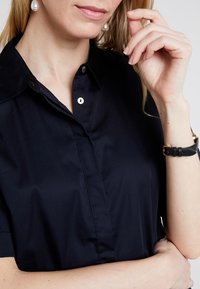 Donna che indossa una camicia nera con colletto, orecchini di perle e un orologio da polso nero, con una mano vicino al viso su uno sfondo semplice.