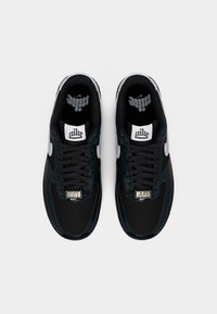 Ζευγάρι μαύρα αθλητικά παπούτσια Nike Air Force 1 με λευκά σήματα swoosh, μαύρα κορδόνια και μεταλλικές ετικέτες «AF1» σε λευκό φόντο.