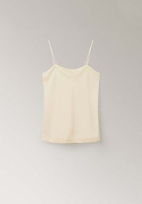 Camisole sans manches de couleur crème avec fines bretelles spaghetti suspendue sur un fond uni gris clair.