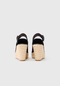 Ζευγάρι μαύρα σανδάλια πλατφόρμας με τακούνια από πλεγμένο μπεζ σχοινί και λουράκια στον αστράγαλο, φωτογραφημένα από πίσω σε απλό ανοιχτόχρωμο φόντο.