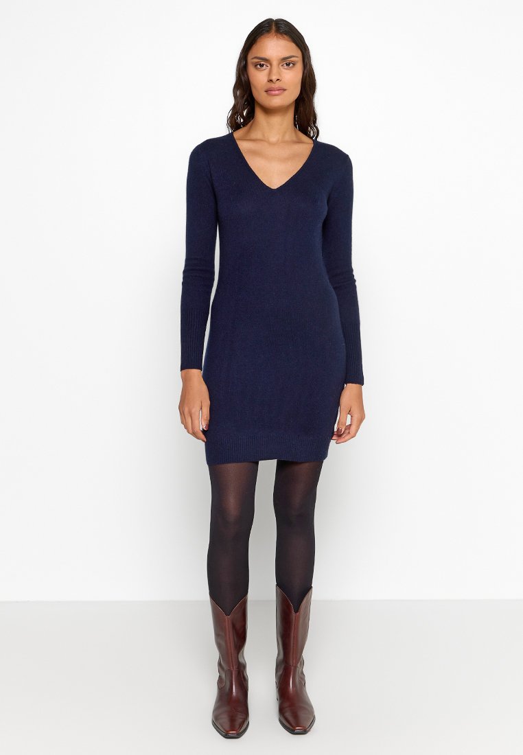 Marineblaues Strickkleid mit langen Ärmeln und V-Ausschnitt. Figurnahes Silhouette, bis zur Mitte des Oberschenkels reichend, kombiniert mit schwarzen Strumpfhosen und braunen Absatzstiefeln.