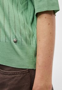 Πλεκτό μπλουζάκι με πράσινες ρίγες και κοντά μανίκια, με ribbed τελείωμα και λεπτομέρεια από ασημένιο κουμπί κοντά στην κάτω πλευρά.