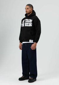 Zwarte hoodie met witte tekst "HE TRAINS FOR BOXING," gecombineerd met losse marineblauwe spijkerbroek en chunky zwarte schoenen met contrasterende veters.