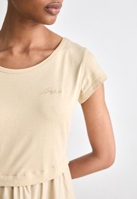 Beige t-shirt met korte mouwen en een ronde halslijn, voorzien van een delicate geborduurde logo op de borst. Gladde textuur, slim fit silhouet, zichtbare naden.