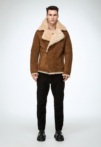 Braune Shearling-Jacke mit breitem cremefarbenem Kragen und Manschetten, ausgestattet mit Reißverschluss, kombiniert mit schwarzen Hosen und klobigen schwarzen Stiefeln.