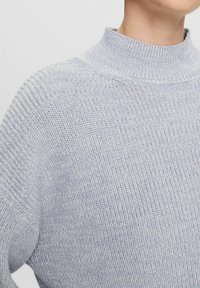 Lichtgrijze gebreide trui met een hoge kraag, voorzien van een gestructureerd patroon en geribbelde manchetten. De stof oogt zacht en comfortabel.