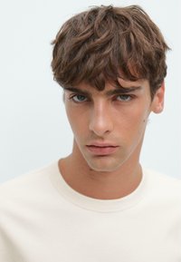 Portret van een jonge man met kort, warrig bruin haar, gekleed in een licht crèmekleurig t-shirt. Neutrale achtergrond versterkt de aandacht voor de gelaatstrekken.