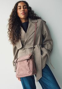 Beige raksts mētelis ar jostu, saskaņots ar rozā ādas crossbody somu. Zilas plaša piegriezuma džinsus, stāv uz gaiši pelēka fona.