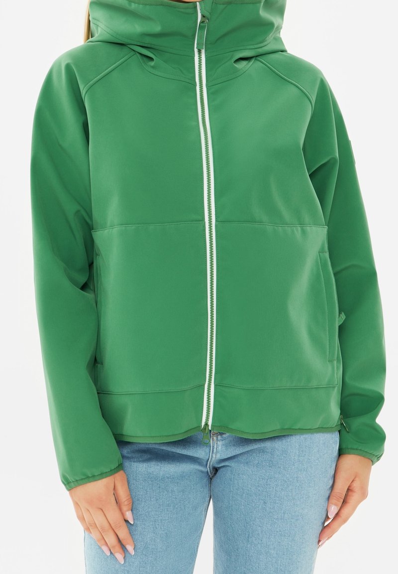 Personne portant une veste à capuche zippée verte et un jean bleu clair, les mains détendues le long du corps sur un fond blanc.