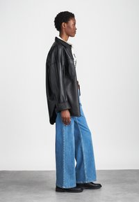 Μαύρο δερμάτινο oversized μπουφάν, λευκό πουκάμισο από κάτω, ανοιχτό μπλε φαρδύ παντελόνι και μαύρες μποτάκια μέχρι τον αστράγαλο; το μοντέλο στέκεται σε προφίλ απέναν�τι από έναν μονόχρωμο φόντο.