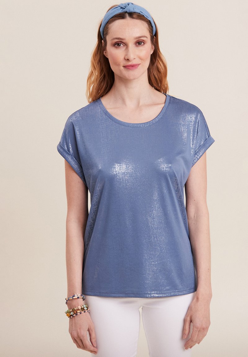 Blauw T-shirt met korte mouwen en een glanzende textuur en opgerolde manchetten. Heeft een ronde halslijn en een ontspannen pasvorm, gedragen met witte broeken en kleurrijke armbanden.