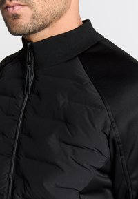 Zwarte jas met ritssluiting, geribde kraag en een gewatteerde structuur aan de voorkant, met gladde stof op de schouders.