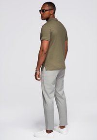 Polo shirt verde oliva con maniche corte, pantaloni grigi su misura e sneakers bianche. Gli accessori includono occhiali da sole scuri e un orologio d'argento.