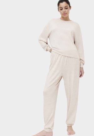 Kvinde stående barfodet iført matchende beige langærmet top og løse bukser mod en hvid baggrund.