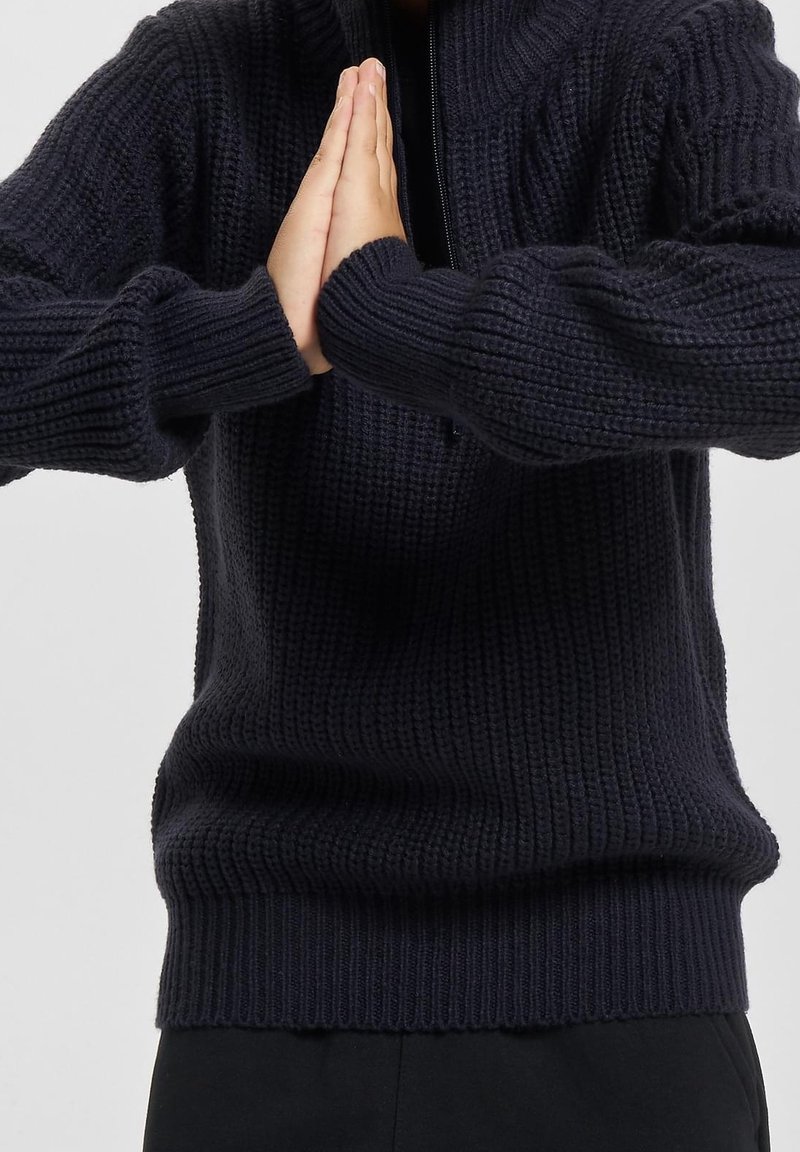Personne portant un pull en laine foncé avec des manches couvrant les poignets, les mains pressées l'une contre l'autre dans un geste de prière ou de salutation.