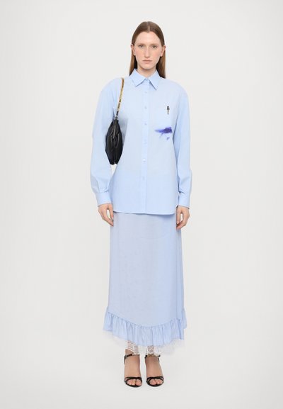 Camicia a maniche lunghe blu chiaro con bottoni e tasca; indossata sopra una gonna blu chiaro coordinata con pizzo. Borsa nera.