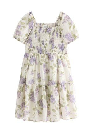 Lachend jong meisje staat met één been vooruit, draagt een bloemenjurk, een witte gebreide cardigan, lavendelkleurige sokken, beige schoenen en een paarse haarstrik.