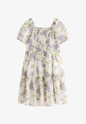 Smilende ung pige står med det ene ben fremad iført en blomsterkjole, hvid strikket cardigan, lavendelfarvede sokker, beige sko og en lilla hårsløjfe.