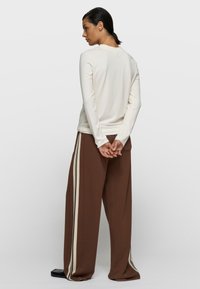 Camisa crema de manga larga con cuello redondo, combinada con pantalones marrones sueltos con rayas laterales crema y negras. Textura de tela suave.