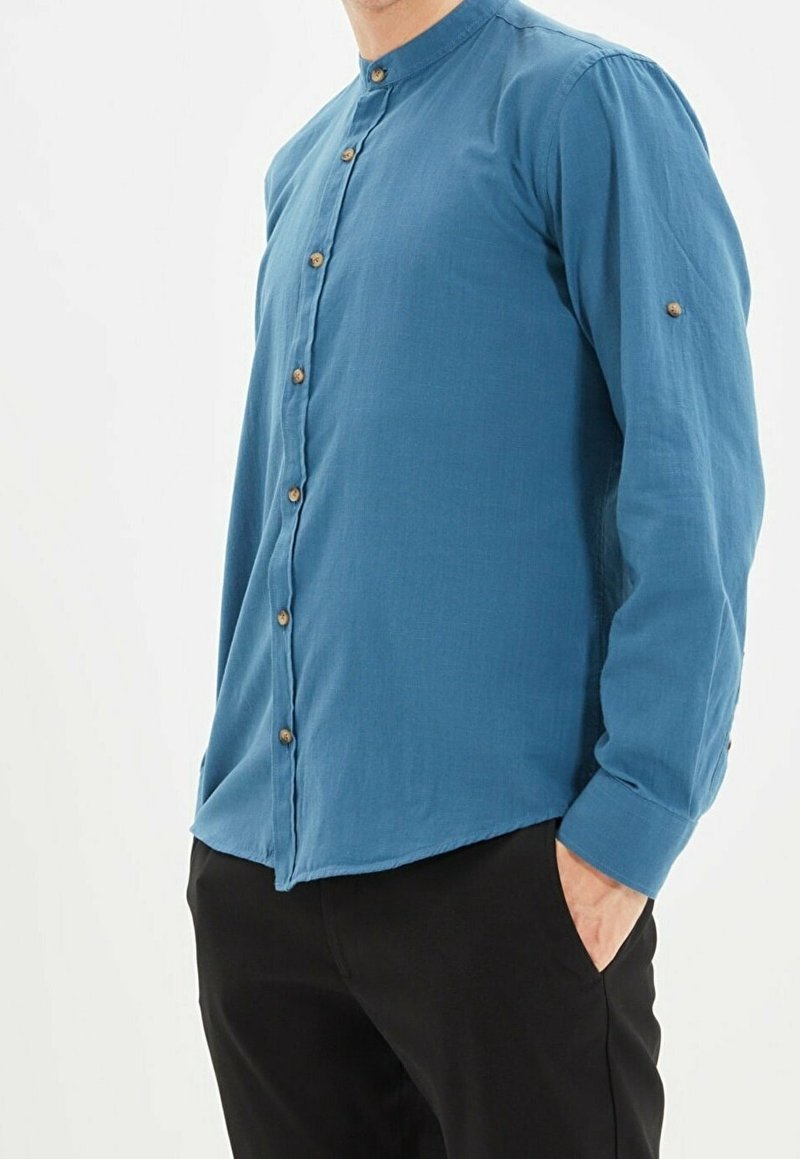 Homme portant une chemise bleue à col mandarin avec des boutons en bois et un pantalon noir, une main posée décontractée dans la poche, sur un fond clair uni.