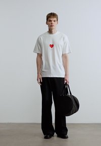 Λευκό βαμβακερό μπλουζάκι με κόκκινη καρδιά, συνδυασμένο με μαύρο παντελόνι φαρδιάς γραμμής. Κρατώντας μαύρη στρογγυλή τσάντα, στέκεται σε ουδέτερο φόντο.