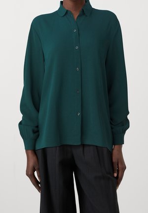 Personne portant une chemise boutonnée à manches longues vert foncé avec col et un pantalon noir, debout devant un fond clair uni.