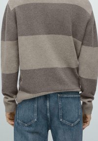 Pletený svetr se širokými šedými a béžovými pruhy, žebrovanými manžetami a lemem, doplněný džínami střední modré barvy s zadními kapsami.